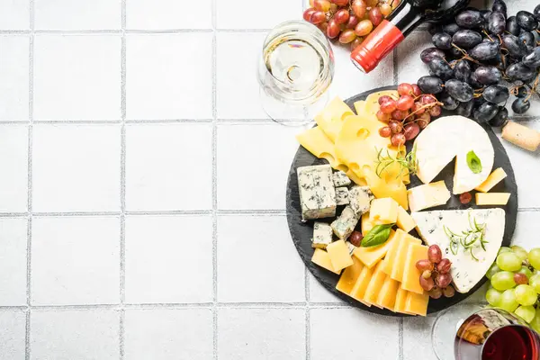 Cheese platter with craft cheese assortment on slate board at white tile background. Top view.