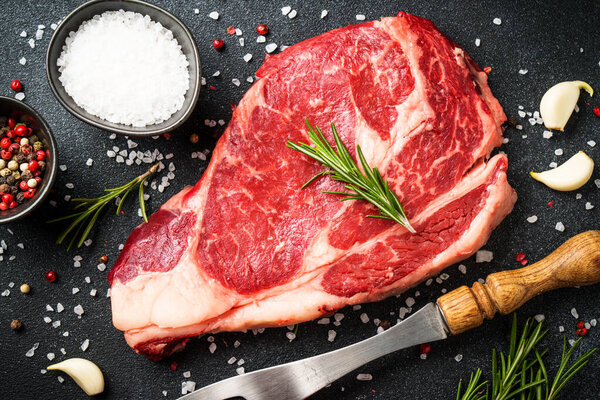 Beef steak with spices on black background. Ribeye steak raw meat. Top view with copy space.
