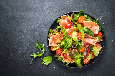 Siyah arka planda yeşil salata. Jamonlu taze salata, yeşil salata yaprakları ve domates. Metin için boşluk olan üst görünüm.