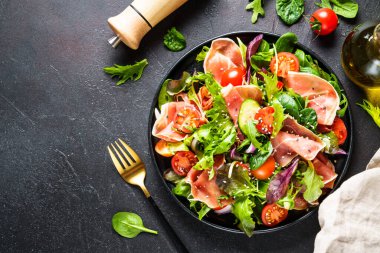 Siyah arka planda yeşil salata. Yeşil salata yaprakları, tütsülenmiş et ve domates. Metin için boşluk olan üst görünüm.