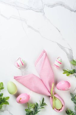 Tavşan kulaklı Paskalya kompozisyonu, beyaz arka planda renkli yumurtalar ve yeşil bitkiler.