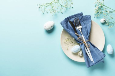 Mavi üzerine Paskalya masası. Beyaz tabak, çatal bıçak, Paskalya yumurtası ve çiçekler..