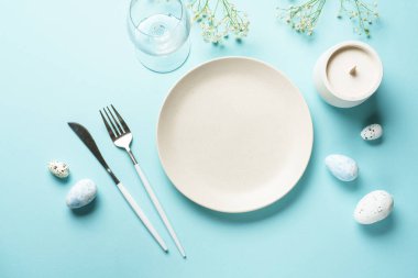 Mavi üzerine Paskalya masası. Boş tabak, çatal bıçak takımı, paskalya yumurtası ve çiçekler. Düz görünüm.