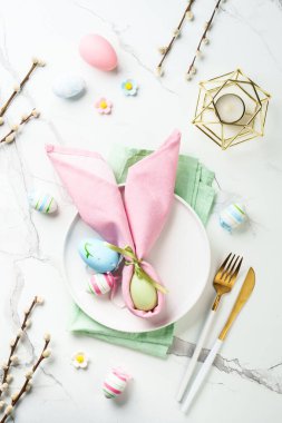 Bahar masası, beyaz tabak, altın çatal bıçak takımı ve renkli paskalya yumurtaları. Kopyalama alanı olan üst görünüm.