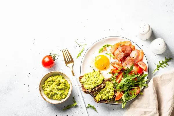 Healthy breakfast or lunch. Beacon, eggs, toast with avocado and fresh salad. Top view on white kitchen table.
