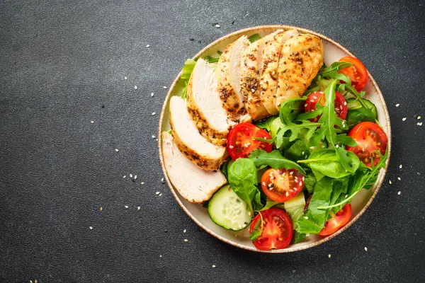 Siyah arka planda tavuk salatası. Izgara tavuk göğsü, domates ve yeşil yapraklar. Tasarım için alanı olan üst görünüm.