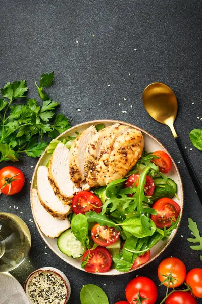 Siyah masada ızgara tavuk eti ve yeşil salata. Arka planda yemek malzemeleri var. Kopyalama alanı olan üst görünüm.