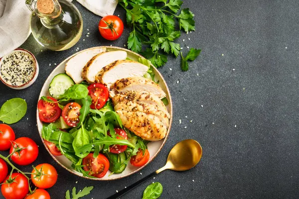 Siyah masada ızgara tavuk eti ve yeşil salata. Arka planda yemek malzemeleri var. Kopyalama alanı olan üst görünüm.