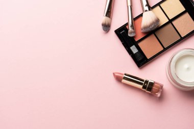 Göz farı paleti, fosforlu kalem, krem, ruj ve kozmetik fırçalar. Pembe arka planda düz uzandı.