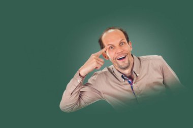 Genç adam parmağını şakağına dayayıp el kol hareketi yapıyor. Arka planda yeşil çam gölgesinde izole edilmiş.