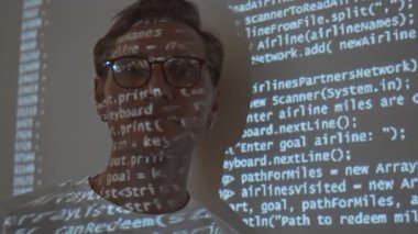Medium closeup portrait of young Caucasian man in eyeglasses looking at camera with program code projected on his face