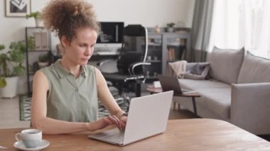 Kadın programcının bilgisayarda yazarken ve evde çalışırken sabah kahvesini yudumlarken orta boy PAN görüntüsü.