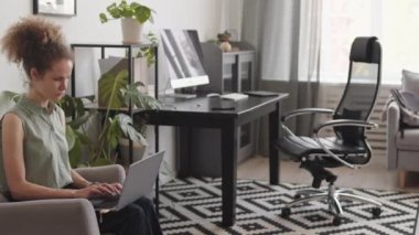 Genç beyaz kadının ev ofisinde çalışırken dizüstü bilgisayar kullanmasının ardından masa başında oturup bilgisayar üzerinde çalışmaya devam etmesinin PAN 'ı
