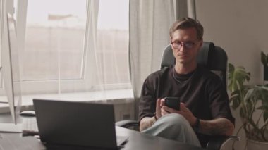 Medium slowmo of tattooed Caucasian male programmer in black tee and eyeglasses scrolling on smartphone and resting in desk chair after work at home office