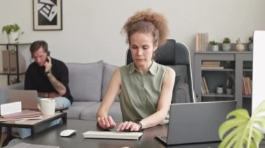 PAN of young Caucasian business couple working in team at home office. Woman typing on laptop while her partner or colleague making phone call sitting on couch in background