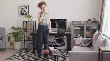 Full length slowmo of female software developer talking on smartphone with colleague while working with program coding on pc computer at home office