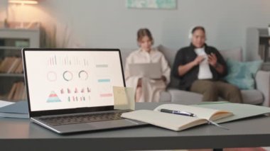 Slowmo of business diagrams on laptop screen at foreground and interracial lesbian couple sitting on sofa at background. One of women working on laptop while her partner listening to music nearby