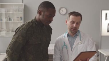 Medium slowmo of Black military officer in uniform listening to Caucasian radiologist in white lab coat talking about patients xray showing images on tablet pc, having appointment at modern office