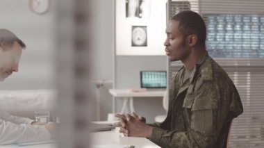 Belden yukarısında, üniformalı Afrikalı Amerikan subayının modern klinikteki sağlık sorunları hakkında doktorla konuşurken yavaşlaması.