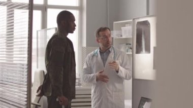 Medium slowmo of African American army officer with walking stick having appointment at radiologist office. Doctor pointing at patients x rays on projector and telling diagnosis