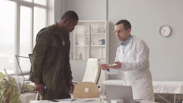 白人男性医師の中低速歩行棒にもたれてアフリカ系アメリカ人兵士に処方を与えます 現代の医師事務所での医療任命を持っています — ストック動画