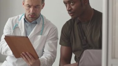 Tilt up slowmo of African American soldier with broken arm listening to Caucasian male doctor holding tablet pc in hands, sitting together on medical couch at doctors office