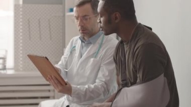 Medium slowmo of injured African American army officer with arm sling and Caucasian male doctor with tablet pc talking during appointment in modern clinic