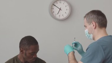 Medium slowmo of brutal African American army officer in khaki being vaccinated against covid by male nurse in face mask