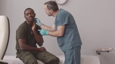 Medium slowmo of Caucasian male medical worker examining neck of African American army officer sitting on couch at doctors office