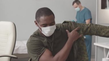 Medium slowmo of African American soldier in face mask taking off his military jacket and being vaccinated by male nurse at modern doctors office