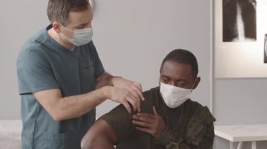 Kafkas erkek askeri doktorunun omzu çıkmış Afrikalı Amerikan askerini muayene etmesi, hastaların röntgenleri arka planda.