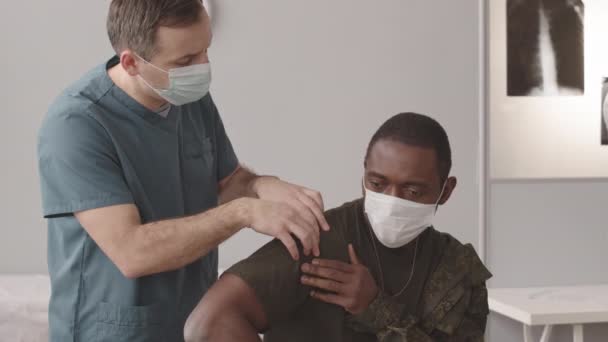 肩の脱臼で負傷したアフリカ系アメリカ人兵士を検査する白人男性軍医の中程度の遅さ 背景にプロジェクター上の患者X線 — ストック動画