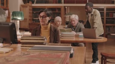 Medium slowmo of mature female librarian in eyeglasses sorting and classifying books at workplace while senior professor and his two diverse university students studying at desk in background