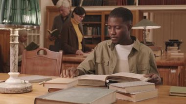 Waist up slowmo of intelligent African American male student reading thick book while studying at university library