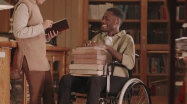 Tekerlekli sandalyede oturan, kucağında kitap yığını olan, başörtüsü ve gözlüklü genç Müslüman bir kadınla konuşan ya da çalışan Afrikalı Amerikalı bir adamın orta yavaşlığı.