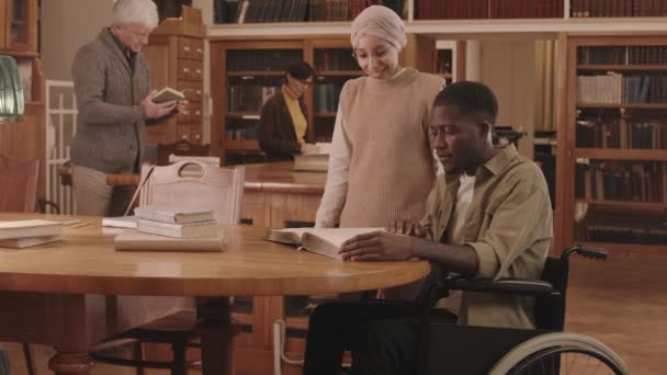 戴着眼镜和头巾的穆斯林女图书管理员与坐在轮椅上的非洲裔美国大学生交谈的速度中等 她们坐在木制书桌边看书 — 图库视频影像