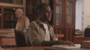 Medium slowmo of blind African American young man reading Braille book sitting by wooden desk at library