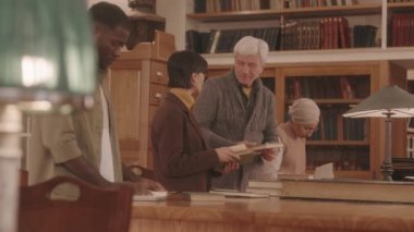 Medium slowmo of Caucasian senior man talking to mature woman in eyeglasses standing by wooden desk in library while other people reading nearby