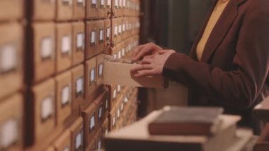 Cropped slowmo of unrecognizable female librarian opening library card index to find some information, looking through cards at wooden drawer