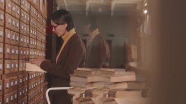 Medium slowmo of mature Caucasian female librarian in eyeglasses standing by drawers with library paper database or archive searching for necessary book