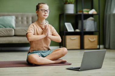 Full length portrait of cute girl with down syndrome doing yoga at home, sitting on mat in lotus position and meditating