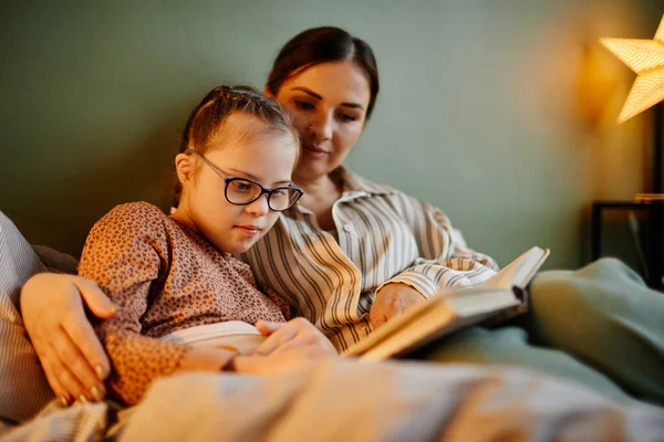 Portrait of little girl with down syndrome reading book at bedtime and relaxing with caring mother embracing her
