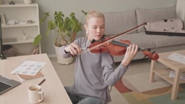 Evde keman çalan, pencerenin önünde masa başında oturan, konsantre olmuş, genç, beyaz bir kadın. Her yerde müzik sayfaları var.