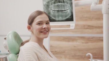 Çağdaş dişçi muayenehanesinde dişçi koltuğunda kameraya gülümseyen ve arka planda diş röntgeni olan genç, beyaz bir kadının belden yukarı portresi.