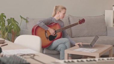 Dizüstü bilgisayarda gitar dersi alan, evdeki koltukta elinde enstrümanla oturan genç bayan müzisyen.