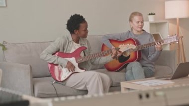 Çok ırklı iki genç kız arkadaşın birlikte akustik ve elektro gitar çalması, gri koltukta oturması.