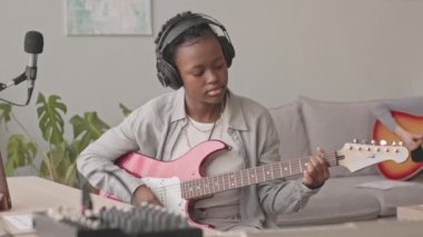 Evde müzik stüdyosunda şarkı söylerken ve arka planda akustik gitar çalarken, kulaklıklı Afrikalı Amerikalı genç bir kadının mikrofonda gitar çalmasının orta yavaşlığı.