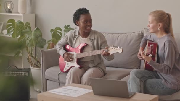 两个有灵感的多民族年轻女子一起弹奏电吉他和声学吉他 坐在舒适客厅的灰色沙发上 速度中等 — 图库视频影像