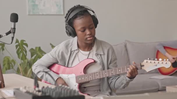 中程度の低速アフリカ系アメリカ人の若い女性のヘッドフォンでマイクでエレキギターを演奏しながら 自宅の音楽スタジオで歌を録音し 彼女の同僚はバックグラウンドでアコースティックギターを演奏 — ストック動画