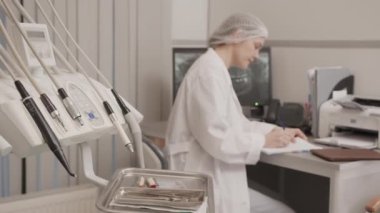 Beyaz laboratuvar önlüklü, tek kullanımlık tıbbi şapkalı, modern diş muayenehanesinde oturan ve fotokopi defterinde hasta dişlerinin röntgenine bilgisayar monitöründen bakan orta derecede yavaş genç bir dişçi.